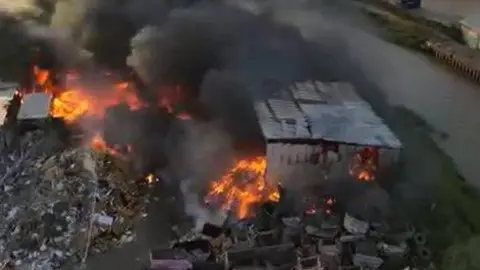 Michael Myers An aerial picture of a scrapyard business on fire. There are orange flames coming from piles of burning rubbish and large black smoke billowing into the air.