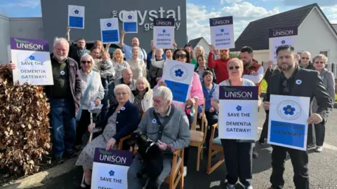 A group of people are stood/sat outside the crystal gateway centre in Amblecote. Many are holding placards which read 'save the dementia gateway.'