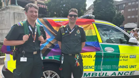 Two men in green paramedic uniforms, wearing rainbow lanyards, smile in front of a West Midlands Ambulance Service car which has been draped in a rainbow flag. Both men have brown hair, while the one on the right wears dark glasses and has rainbow face paint on both cheeks. They are in a city square, with the plinth of a statue on their left and restaurants and bars behind.
