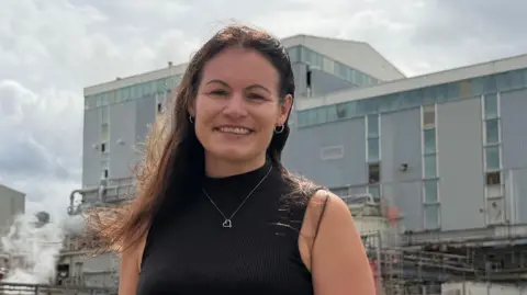 BBC / Mark Denten Karen Askwith is standing in front of a grey industrial building and smiling brightly at the camera. She has long, dark hair and is wearing a black vest.
