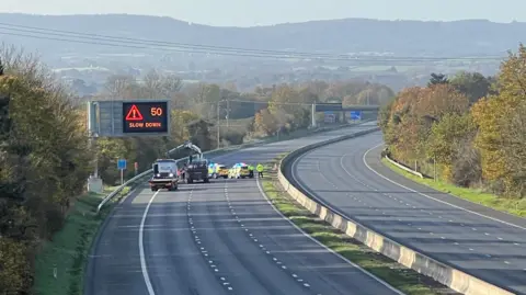 A closed motorway with two police cars and a regular car parked near the hard shoulder. A large digital sign with orange text on a black background says "50 - slow down" with an exclamation mark inside a triangle