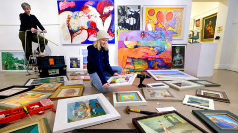 A woman sits on the floor of an art gallery, surrounded by paintings and artwork. Another woman is standing on a small stepladder in the background