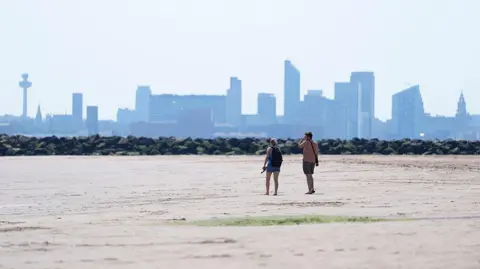 Two people walking on New Brighton Beach on The Wirral, Merseyside. The Liverpool waterfront and skyline is in the distance.