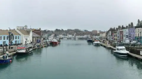 Panoramic image was created using a iPhone.) People walk around the harbour which has boats in the water at Weymouth. 