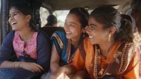 Three women are smiling in a car looking out the window