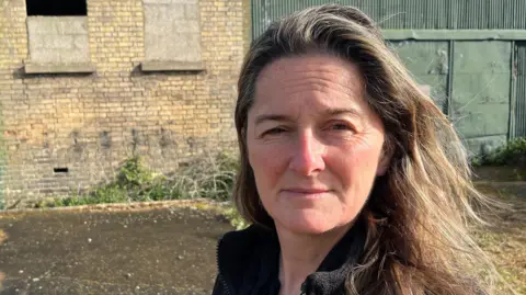 Hayley Dolbear has long brown hair and is wearing a black jacket. She is standing in front of a derelict building which has its windows bordered up and a green shed attached on the right side.