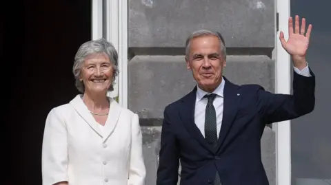 Carney, a man with a navy suit and green tie waving while standing beside Connolly, a woman in a white coat standing smiling.