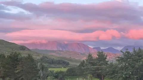Lisa Blackett Large pink clouds hang over mountains in the distance. Closer to the viewer are green fields and hills and trees.