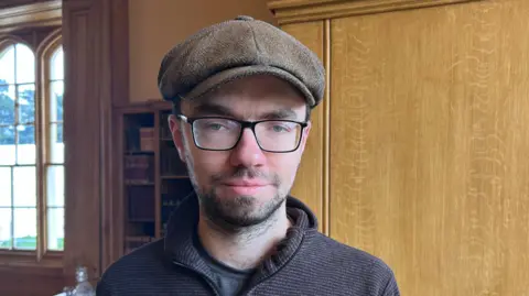 BBC / Kate Bradbrook Man in his late twenties with glasses and a flat cap stands in front of oak panelling and a large window on the right