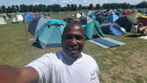 Handout A man in a white shirt taking a selfie. There are a row of tents in the background.