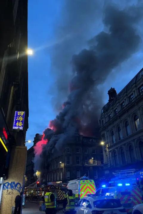 Joe Diaz A city centre street with police officers and police cars with blue sirens outside a huge building with black smoke filling the blue evening sky