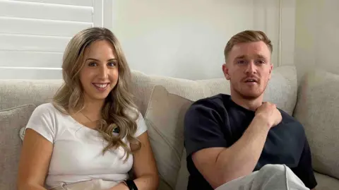 Jack Sparkes and partner Abbie Denton are sitting on a beige sofa looking at the camera. She has long, curled blonde hair and is wearing a white T-shirt. He has short, blonde hair and stubble on his face, he is wearing a black T-shirt.