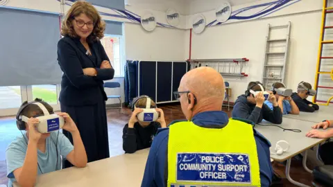 Policing Minister Diana Johnson watches on as schoolchildren at St Gregory's Primary School in Coventry take part in a virtual reality knife crime session run by West Midlands Police. The children are wearing white VR headsets which cover their eyes. A Police Community Support Officer has his back to the camera while he instructs wearing a yellow high visibility vest.
