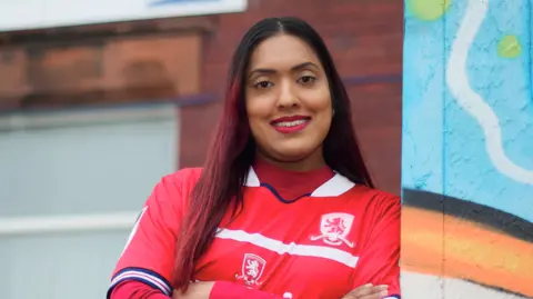 Tom Banks and Paul Thompson Sahida Ditta is leaning against a brightly painted pillar in a red and white Middlesbrough shirt. She has long black hair which has been dyed red towards the ends and has her arms folded. She is smiling broadly.