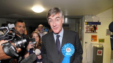 Jacob Rees-Mogg dressed in a suit, with a blue conservative rosette. He is looking straight into the camera, while journalists and photographers follow next to him.