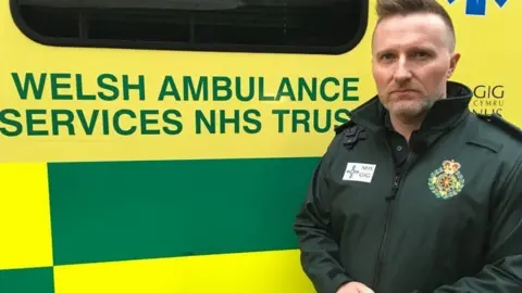 Jason Killens, kepala layanan Ambulans Welsh, berseragam berdiri di dekat salah satu kendaraannya