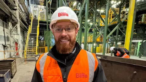 A man with a ginger beard is wearing a white hard helmet that says 'Biffa' and a black jacket. He is also wearing an orange high vis jacket and is standing in an industrial factory.