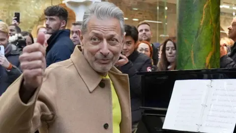 Actor Jeff Goldblum with a thumbs up while sat at a piano in St Pancras station