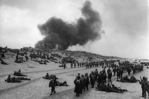 Hulton-Deutsch Collection/CORBIS/Corbis via Getty Images Hundreds of men and boys in military uniform standing, walking and sitting on the beach. There is a plume of black smoke in the background. It is a black and white image.