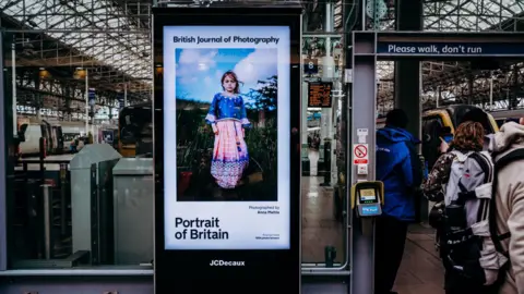 Anna Mehta A portrait of a girl in an Indian dress standing in a rural area is shown on a train station advertising board.