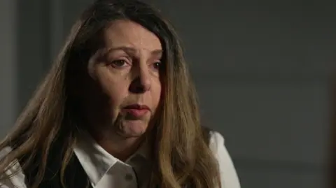 A woman in black and white shirt with light brown hair looks towards a reporter off camera. 