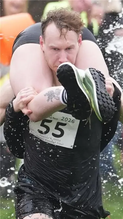 A man with a marathon number on carrying a woman over his shoulders.