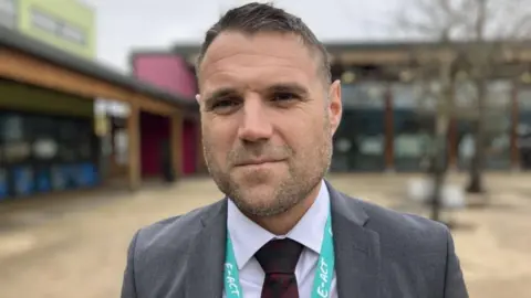 Headteacher Guy Swallow stood outside Merchants' Academy Secondary School. He is wearing a grey suit, white shirt and a tie. The school building behind him is blurred. 
