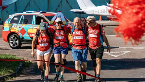 Sophy Dunn, Helene Poursain, Duncan Bootland and Warren Porritt in red KSS shirts walking with backpacks on and their arms around each other. They are walking towards a red ribbon. A KSS vehicle and the tail of a light aircraft can be seen behind them.