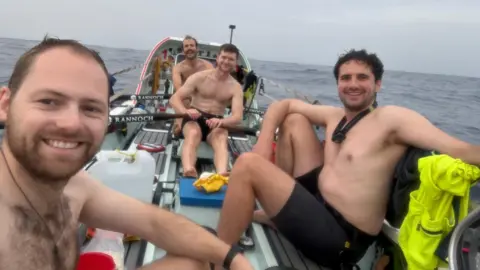 Atlantic Sea Donkeys Four topless men in a rowing boat smile at the camera with the sea behind.