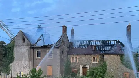 Robin Legg A burnt roof of the listed school building with fire hoses shooting at it.