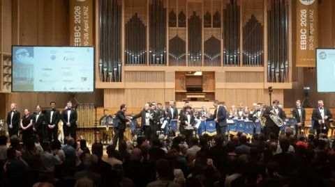 Charlie Chan In the foreground an audience sits in relative darkness. Audience members are clapping. A stage is lit up in the background and a band are seen on their feet all facing the audience. Most of the players are wearing black suits with white shirts. In the background, at the back of the stage, are silver organ pipes of a large organ which takes up much of the backdrop. There is a large TV screen at either side of the stage showing competition branding and sponsor logos.