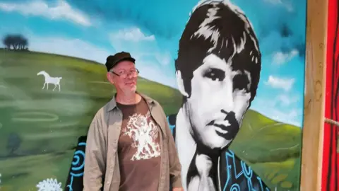 Jack Sant Artist Paul Boswell wearing glasses and a black cap standing in front of the large Reg Presley mural in the pub with the white chalk horse on the hills and UFO in the background.