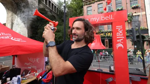 North News Joe Wicks, who has swept back long dark hair, a close cropped dark beard and wearing a short sleeved black t-shirt. He is holding some kind of klaxon and behind him is a red bell hanging from a red stand.