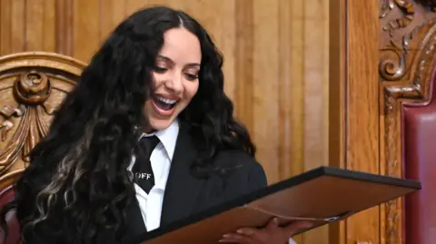 South Tyneside Council Jade Thirlwall looking down at a picture she has been presented with from South Tyneside Council. She has long black hair and is wearing a black blazer and tie with a white shirt. She has her mouth open wide in a smile.