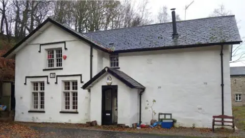 St Robert and St Alice Chapel at Dodding Green. The building is white with black window sills and a slate roof. There is a central doorway under a pitched roof. To its left is a wing that stands proud of the main building and has three windows, two on the ground floor and one centrally above. There is also a small window on the first floor above the roof over the main door. The part of the building to the right of the door is a single white painted wall with no windows.