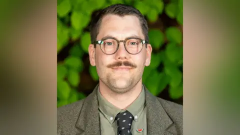 Welwyn Hatfield Borough Council A smiling Max Holloway looking directly into the camera. He has short dark hair and a moustache. He is wearing glasses with circular frames and a olive-coloured suit jacket and shirt, with a black tie that has a pattern.