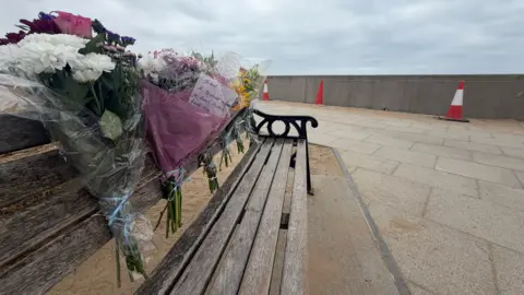 BBC/Jim Scott A side-view photo of a bench at Seaton Carew beach, where flower tributes to Wayne Taylor and Ian Pascoe have been attached with notes.
