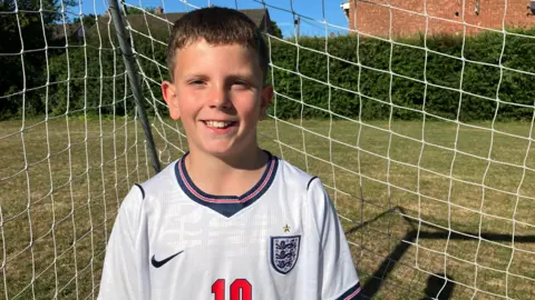 Henry Godfrey-Evans/BBC A child standing in front of a goal with a number 10 England top on. 