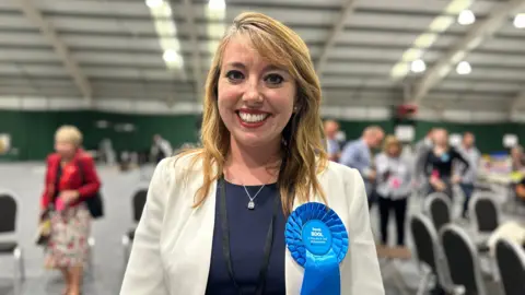 Carroll Weston/BBC A woman in a cream blazer and blue rosette smiles at the camera. She appears to be an an election count.
