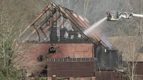 Roof frame of barn building after fire - the roof is gone with just the frame left. Water is being sprayed into the barn from ladder platform by a fire crew.