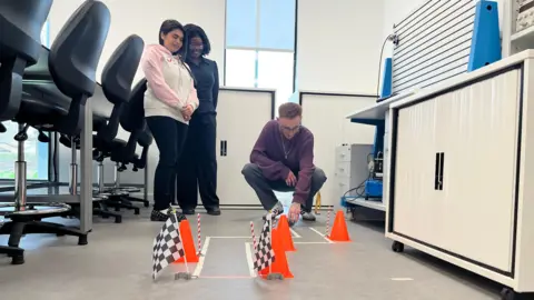 Emma Baugh/BBC Three people next to a course mapped out on the floor with cones and flags, helping an AI ball navigate through.