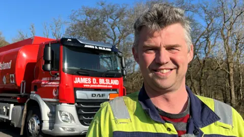 BBC Stuart Youens standing in front of a red lorry, wearing a high vis boiler suit and a striped tshirt.