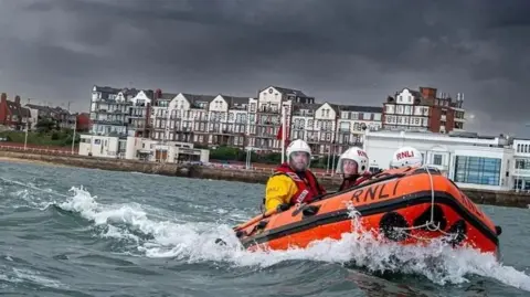 Three lifeboat crew members in RNLI (Royal National Lifeboat Institution) gear riding a bright orange rescue boat on water near a Bridlington town. Stormy dark clouds are overhead and large buildings are in the background.