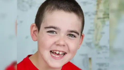 CLC PORTRAITS A boy with dark hair and his mouth open is looking at the camera. He is wearing a red top.