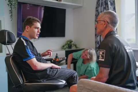 Elliot sits in a chair in his new flat wearing a blue and black sports top and grey jeans. He is looking at his father Jonathan who is sitting near him on a grey sofa wearing a black polo neck with white writing on the back. Mum Chris is sitting on the floor looking up at Elliot with the patterned curtains behind her. She is smiling and wearing a green t shirt. She has blonde shoulder-length hair.