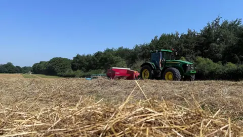 Wiff Straw on the ground and a tractor in the distance and some trees behind that.