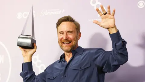 EPA David Guetta holding an American Music Awards trophy which is triangular in shape. He is wearing a longsleeved blue buttoned shirt with pockets on the chest.