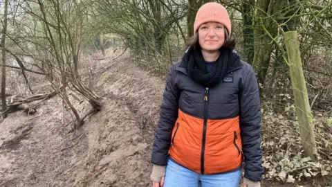 A lady standing on a muddy path, she is wearing a navy and orange coat with blue jeans. She is smiling into the camera and wearing a pink hat. 