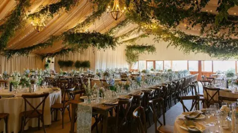 Emma Rajan A wedding tent with green foliage and lights hanging from the ceiling. There are tables made up with flowers and cutlery ready for a wedding