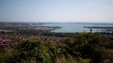 Portsmouth City Council The view from Portsdown Hill, which overlooks Portsmouth there is trees and other greenery in the foreground and housing and the sea in the background